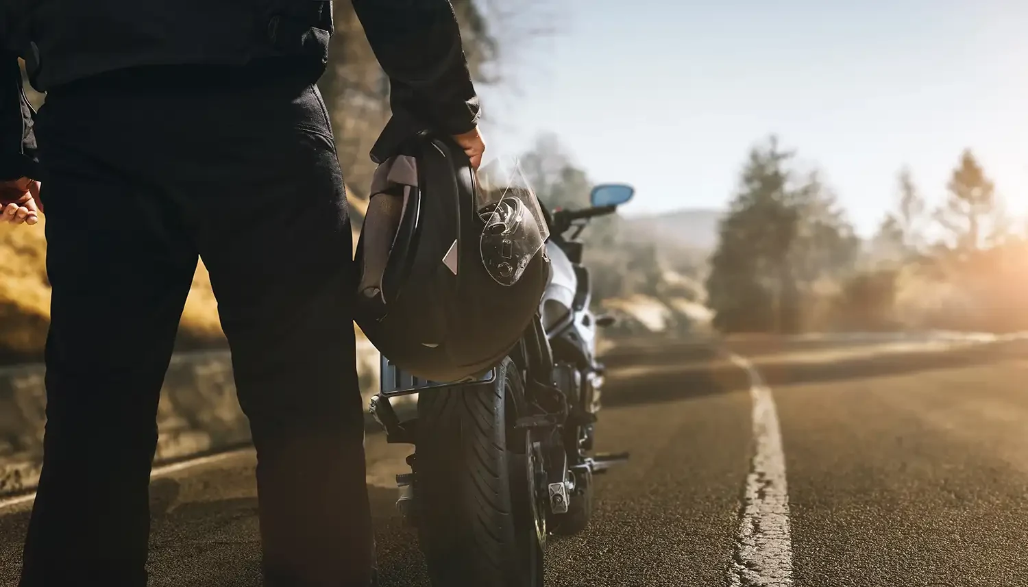 Motorradfahrer steht mit Helm in der Hand neben seinem Motorrad auf einer Landstraße bei Sonnenuntergang, im Hintergrund verschwommene Bäume und Natur.