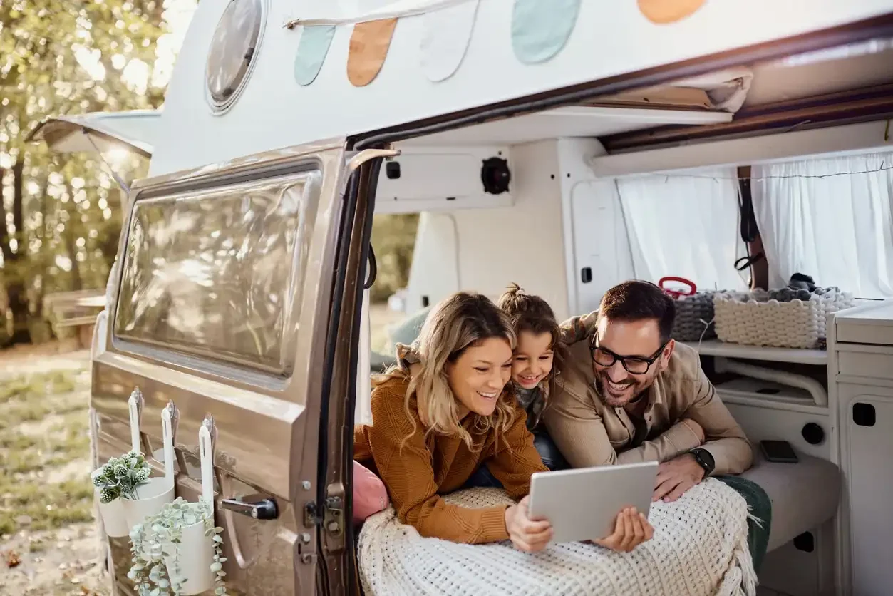 Familie mit Kind liegt lächelnd in einem Wohnmobil und schaut gemeinsam auf ein Tablet, umgeben von gemütlichen Decken und Kissen bei Sonnenschein.