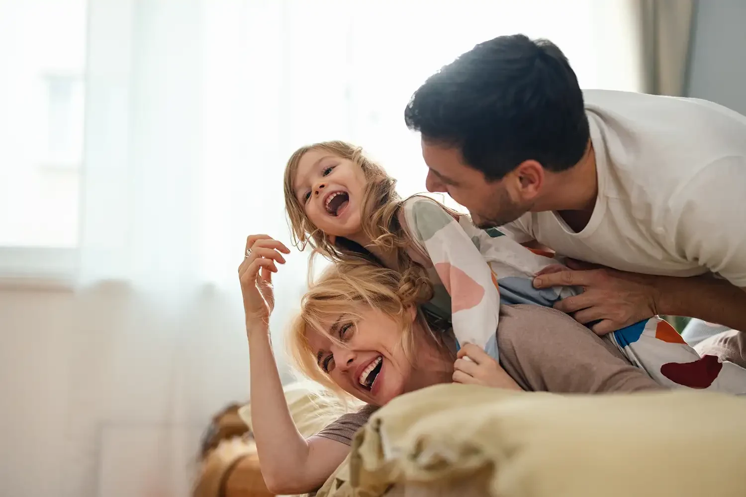 Eine fröhliche Familie spielt zusammen auf einem Sofa - Vater, Mutter und Kind lachen ausgelassen beim gemeinsamen Toben in hellem Wohnzimmer.