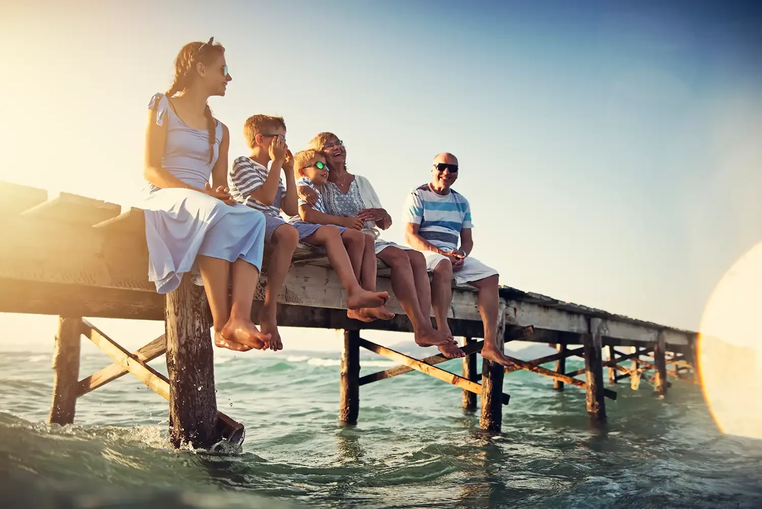 Eine Familie mit fünf Personen sitzt auf einem Holzsteg am Meer bei Sonnenuntergang, alle tragen Sommerkleidung und Sonnenbrillen.