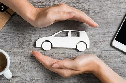 Zwei Hände umschließen schützend ein weißes Auto-Symbol aus Papier auf einem Holztisch, daneben eine Kaffeetasse und ein Smartphone, symbolisierend Kfz-Versicherungsschutz.